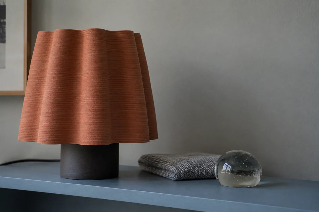 Terracotta-coloured layered lampshade beside folded grey fabric and a glass paperweight.