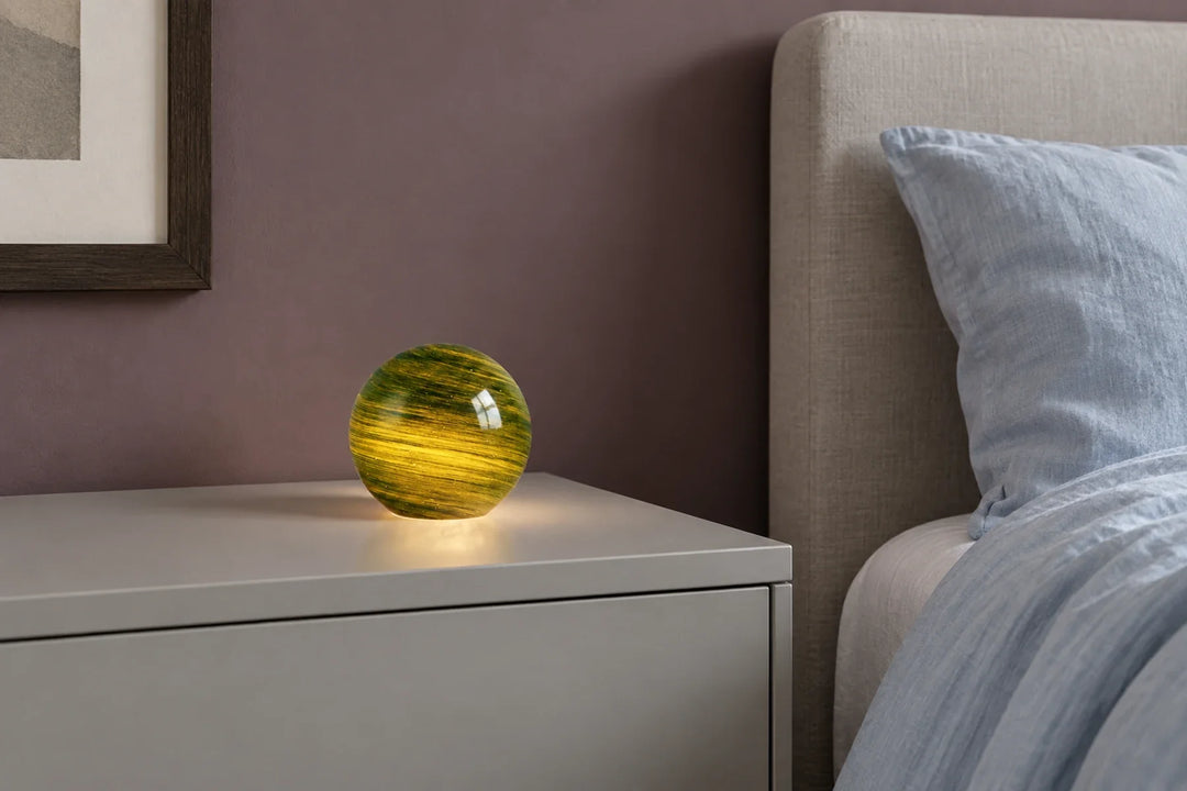 Green and gold spherical glass lamp on a grey bedside cabinet beside blue linen bedding.
