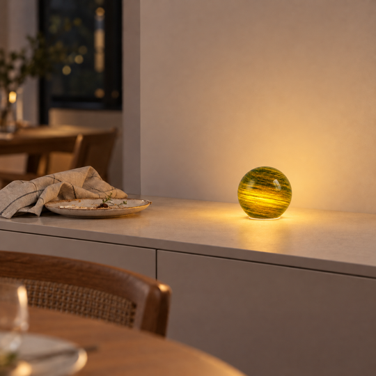 Green and yellow Orbis table lamp glowing on a sideboard beside a dinner plate.