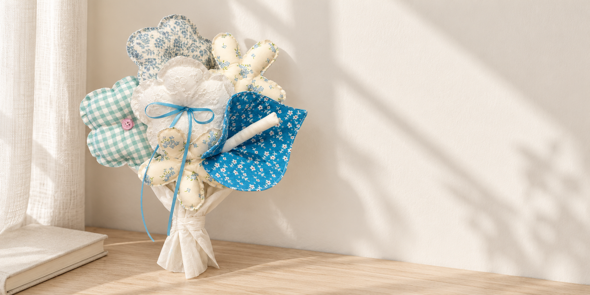 Six patterned fabric flowers with a pink button, ivory lace and a blue ribbon, wrapped on a pale wood table.
